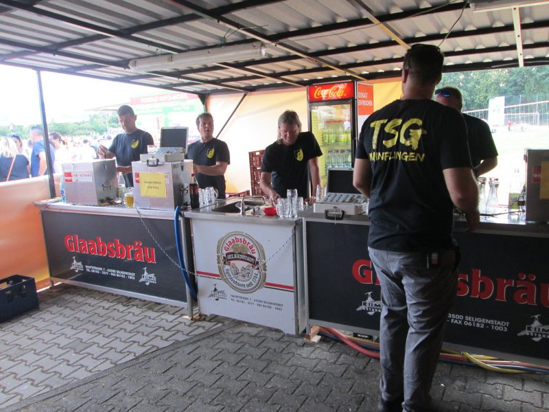 5 Männer in schwarzen Shirts mit gelben Rückenschriftzug TSG Mainflingen hinter der Getränkeausgabe beim Mainpokal 2019