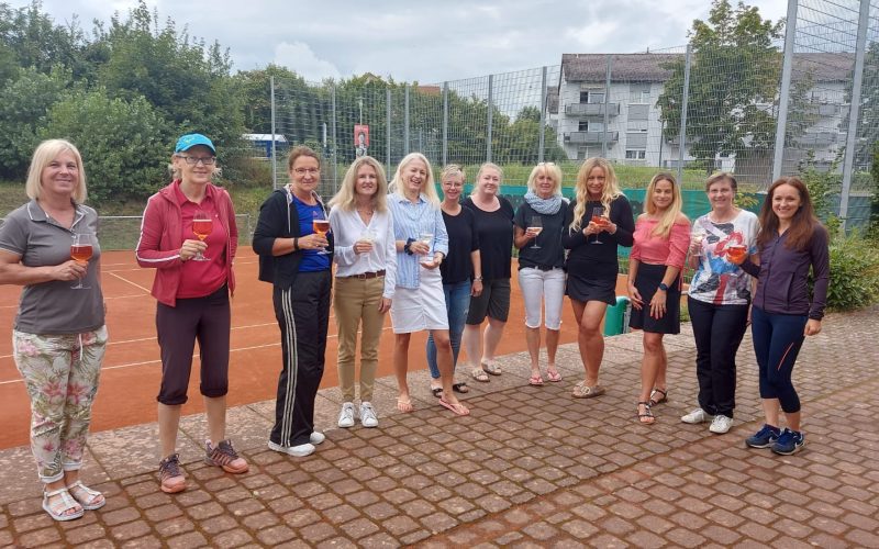 Gruppenfoto. 12 Frauen auf dem Tennisplatz weitestgehend nicht in Sportkleidung. 2/3 hält ein Weinglas in der Hand. Anlass: Frauen Brunch 2021
