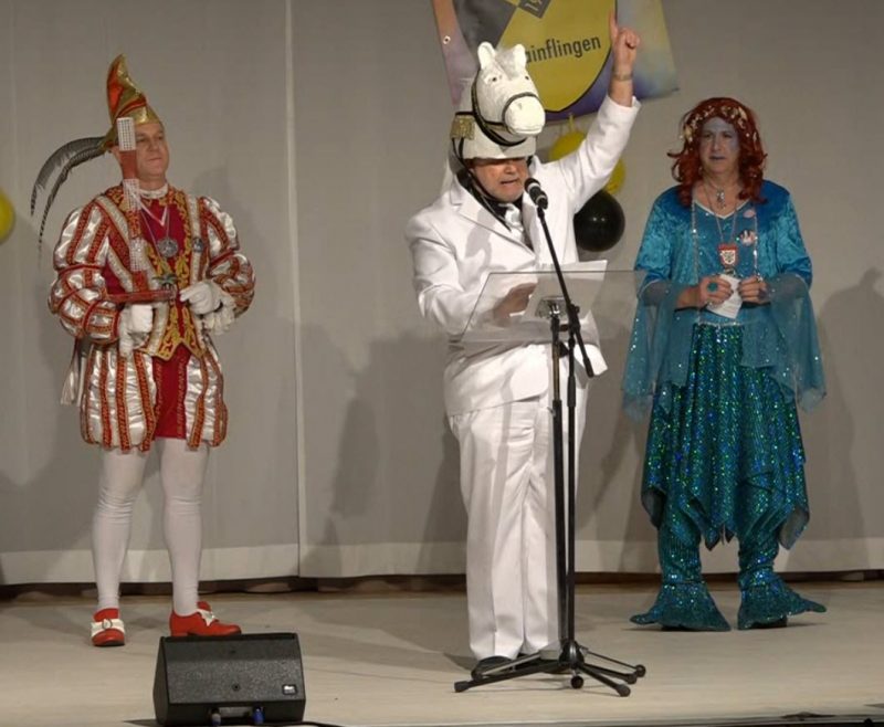Mainflinger Dreigestirn auf der Bühne: Prinz, Schimmel und Meerjungfrau. Alle drei sind Männer. Der Prinz im typischen Kostüm mit weißer Strumpfhose, roten Schnallenschuhen, und einer rot,weiß, silber, goldenen gestreiften Jaclle, weiße Handschuhe und Narrenkappe. Der Schimmel trägt einen weißen Anzug und einen Hut der einen Schimmelkopf darstellt. Die Meerjungfrau trägt einen dunkelblauen Pajettenrock, ein hellblaues Oberteil und eine kupferrote Perrücke mit Muschel-Haarband. Der Schimmel steht am Notenständer und hält eine Rede. Im Hintergrund das einfarbig grauweiße Bühnenbild mit drei TSG Wappen mit Narrenkappe und schwarz-gelbe Ballons und Luftschlangen.
