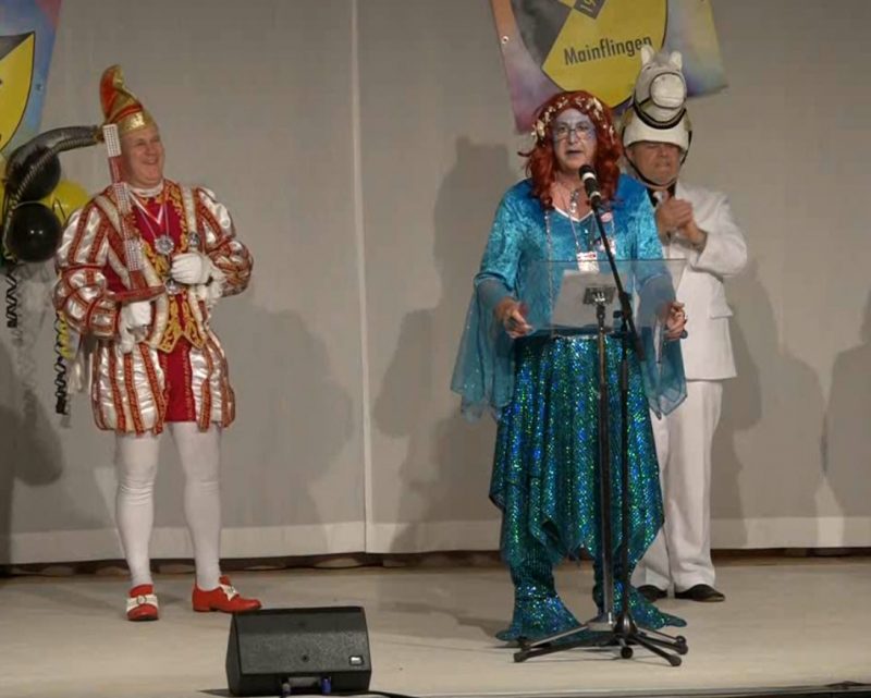 Mainflinger Dreigestirn auf der Bühne: Prinz, Schimmel und Meerjungfrau. Alle drei sind Männer. Der Prinz im typischen Kostüm mit weißer Strumpfhose, roten Schnallenschuhen, und einer rot,weiß, silber, goldenen gestreiften Jaclle, weiße Handschuhe und Narrenkappe. Der Schimmel trägt einen weißen Anzug und einen Hut der einen Schimmelkopf darstellt. Die Meerjungfrau trägt einen dunkelblauen Pajettenrock, ein hellblaues Oberteil und eine kupferrote Perrücke mit Muschel-Haarband. Die Meerjungrau steht am Notenständer und hält eine Rede. Im Hintergrund das einfarbig grauweiße Bühnenbild mit drei TSG Wappen mit Narrenkappe und schwarz-gelbe Ballons und Luftschlangen.