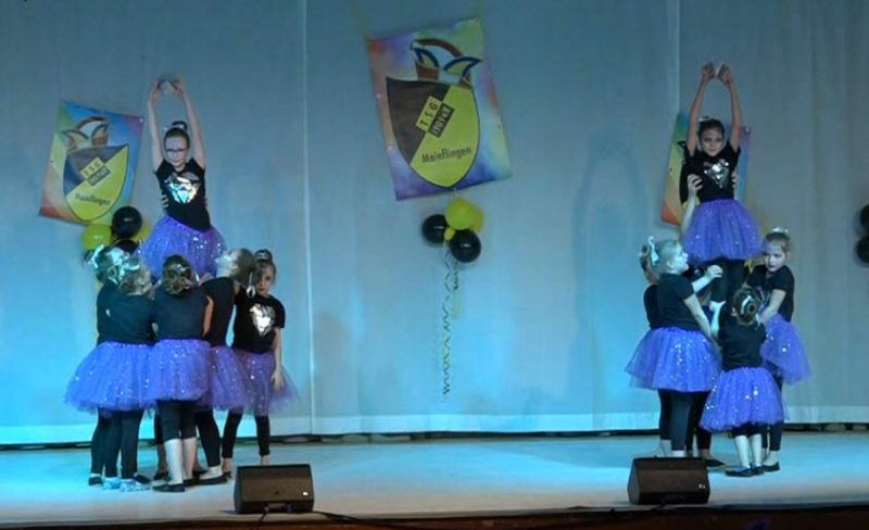 Kindertanzgruppe: Je eine links und rechts am Bildrand. Eine Gruppe von 5-6 Mädchen hält ein Mädchen nach oben. Die Kinder tragen schwarze Oberteile mit einem großen glitzernden Herz auf der Brust und lila Röcke die ebenfalls glitzern. Im Hintergrund der Bühne drei TSG Wappen mit Narrenkappen, sowie schwarz-gelbe Ballons.