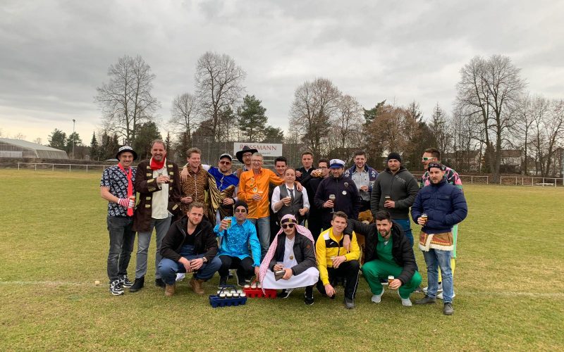 Gruppenfoto auf dem Fußballplatz. 19 Männer, teils verkleidet und die Wirtin des Vereinsheims.
