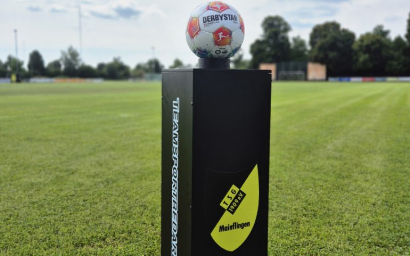 Schwarze eckige Säule 90x30x30 cm, die auf Rasenfläche steht. Auf der Säule liegt ein Derbystar-Fußball. Auf der vorderen Seite der Säule ist das Logo der TSG Mainflingen angebracht. Auf der Seite der Name des Herstellers.