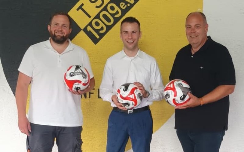 Auf dem Bild v.l.n.r. David Kessler (Vorstand Sportanlage), Tim Spies (SwissLife Select), Dirk Breuhahn (Abteilungsleiter Fußball). Jeder mit einem Fußball in der Hand. Im Hintergrund das TSG Mainflingen Wappen an einer Wand