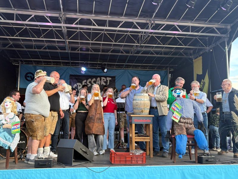 Bieranstich der Mainflinger Kerb 2026. Mehrere Männer und Frauen stehen auf einer Bühne und trinken aus Bierkrügen Bier. Auf der Bühne steht ein Bierfass und afu zwei Stühlen sitzen der Kerbbosch und das Kerbmädel
