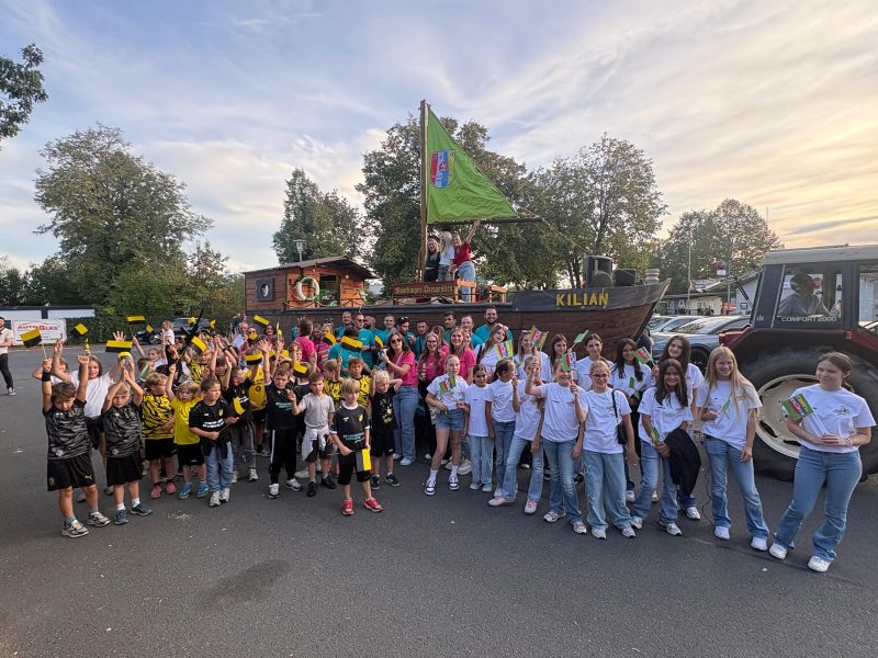Kerbumzug anlässlich der Mainflinger Kerb 2026. Im Hintergrund ein Holzschiff auf einem Anhänger (wie man es vom Fasching kennt), Am Mast weht eine grüne Fahne des Mainflinger Dreigestirns. Vor dem Boot steht eine große Gruppe bestehend aus Kindern in gelb-schwarzen Trikots der TSG Fußballer, Mädchen in weißen Shirt und Männer in türkisen und Frauen in pinken Shirts.